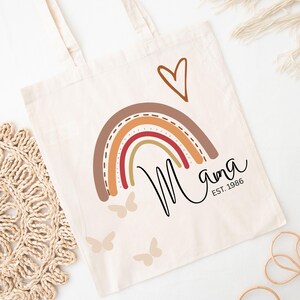 May include: A cream-coloured canvas tote bag featuring a printed design of a multi-coloured rainbow in shades of brown, orange, and red. Above the rainbow is a brown heart outline. The text Mama and EST. 1986 are printed in black, with three small butterflies.