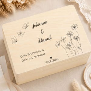 A light wood keepsake box with engraved text and designs, on a cream blanket with dried grasses and a ring on a small plate.
