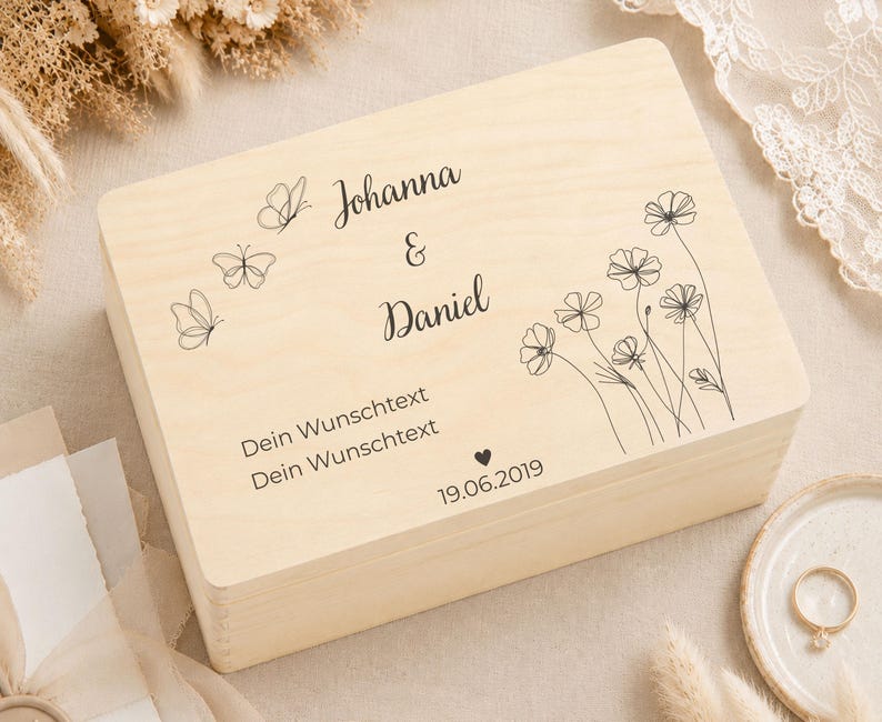 A light wood keepsake box with engraved text and designs, on a cream blanket with dried grasses and a ring on a small plate.