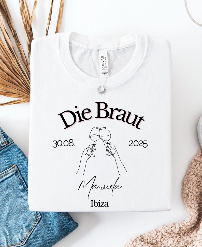 May include: White crew neck sweatshirt with the text "Die Braut" (The Bride) in black script. The design includes a line drawing of two wine glasses clinking, the date 30.08.2025, the name Manuela, and the location Ibiza.