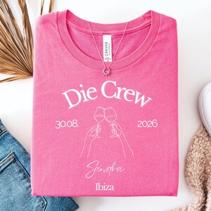 May include: A bright pink t-shirt featuring white screen-printed text that reads Die Crew, 30.08. 2026, Sandra, and Ibiza. The design includes a white line-art graphic of two hands holding wine glasses clinking together in a toast.