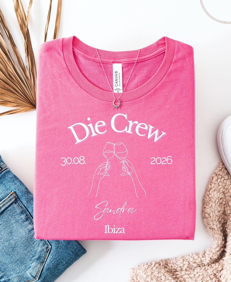 May include: A bright pink t-shirt featuring white screen-printed text that reads Die Crew, 30.08. 2026, Sandra, and Ibiza. The design includes a white line-art graphic of two hands holding wine glasses clinking together in a toast.