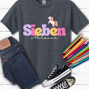 K&ouml;nnte beinhalten: Dunkelgraues T-Shirt mit dem Namen "Sieben Melanie" in einer bunten Schriftart und einer Einhorn-Grafik. Das Bild enth&auml;lt auch eine blaue Jeans, schwarze Turnschuhe und einen Satz Buntstifte.