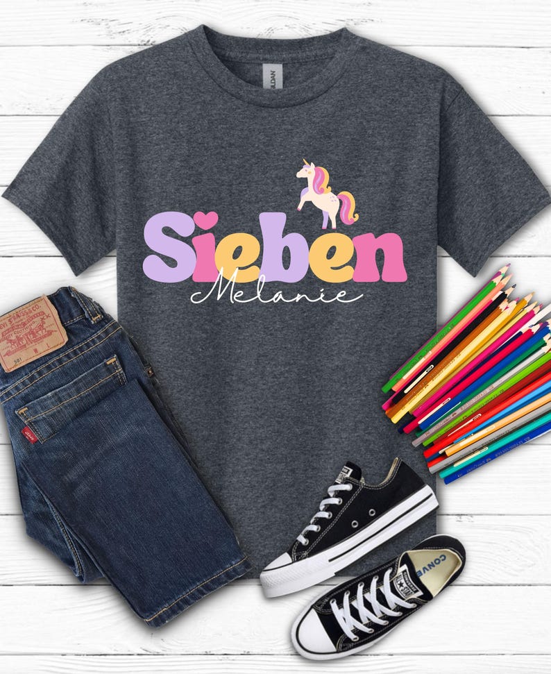K&ouml;nnte beinhalten: Dunkelgraues T-Shirt mit dem Namen "Sieben Melanie" in einer bunten Schriftart und einer Einhorn-Grafik. Das Bild enth&auml;lt auch eine blaue Jeans, schwarze Turnschuhe und einen Satz Buntstifte.