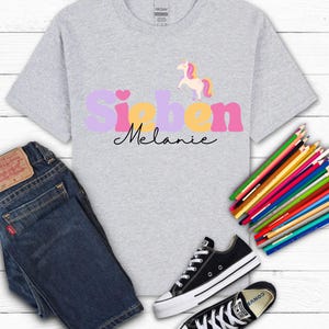 K&ouml;nnte beinhalten: Hellgraues T-Shirt mit dem Namen "Sieben Melanie" in einem farbenfrohen Design, mit einem Einhorn-Motiv. Das Shirt wird mit blauen Jeans und schwarzen Turnschuhen kombiniert. Buntstifte sind seitlich angeordnet.