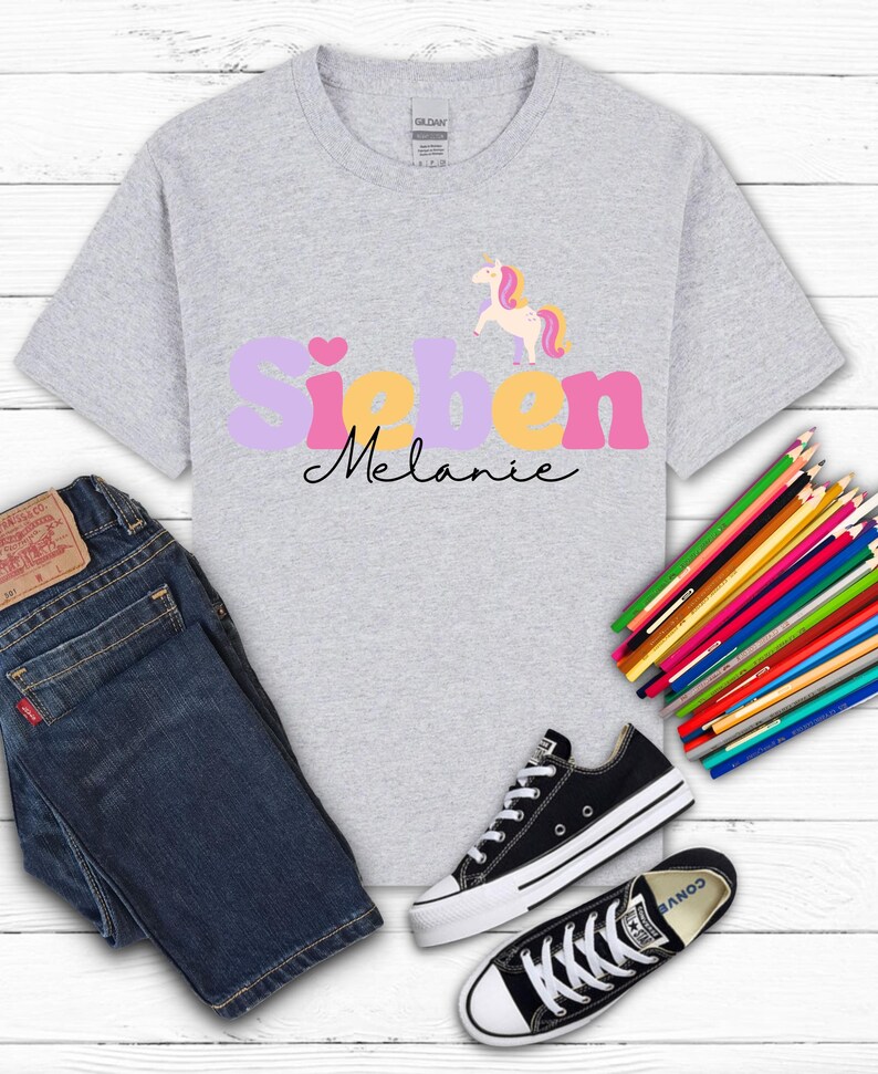 K&ouml;nnte beinhalten: Hellgraues T-Shirt mit dem Namen "Sieben Melanie" in einem farbenfrohen Design, mit einem Einhorn-Motiv. Das Shirt wird mit blauen Jeans und schwarzen Turnschuhen kombiniert. Buntstifte sind seitlich angeordnet.