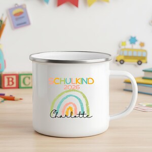 May include: A white enamel mug with a silver rim featuring a colorful graphic of a rainbow and the text "SCHULKIND 2026" in multi-colored letters above the name "Charlotte" written in a black cursive script font on the front of the mug.