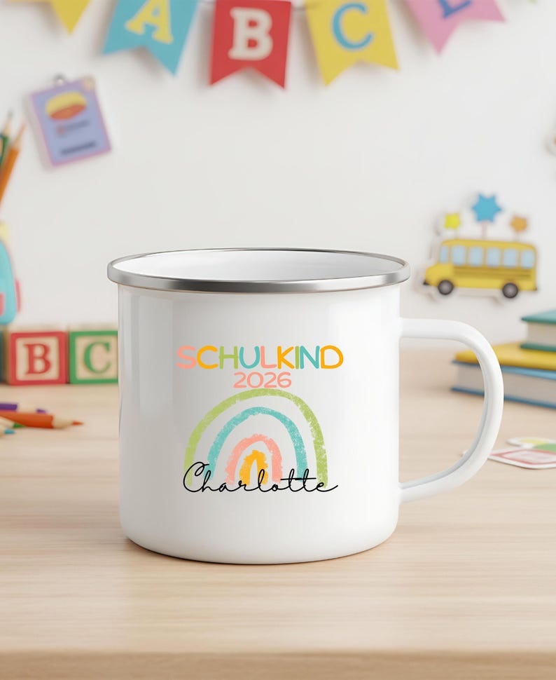 May include: A white enamel mug with a silver rim featuring a colorful graphic of a rainbow and the text "SCHULKIND 2026" in multi-colored letters above the name "Charlotte" written in a black cursive script font on the front of the mug.