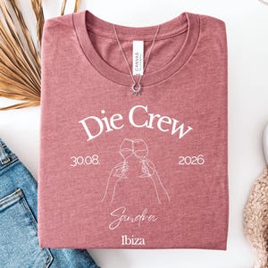 May include: A mauve heather crew neck t-shirt featuring white line art of two hands clinking wine glasses. The text reads Die Crew, 30.08. 2026, Sandra, and Ibiza. A silver sunburst pendant necklace rests on the neckline of the folded garment.
