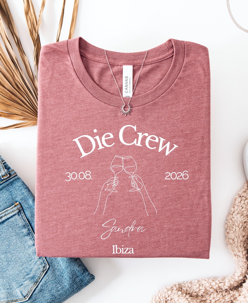 May include: A mauve heather crew neck t-shirt featuring white line art of two hands clinking wine glasses. The text reads Die Crew, 30.08. 2026, Sandra, and Ibiza. A silver sunburst pendant necklace rests on the neckline of the folded garment.