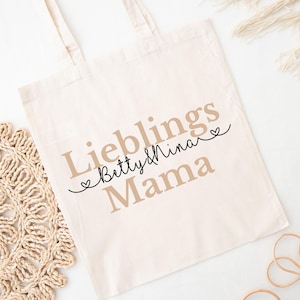 Puede incluir: Bolso tote de color crema con las palabras "Lieblings Mama Betty & Nina" impresas en una mezcla de fuentes y colores. El bolso tiene dos asas y está hecho de una tela suave. El diseño incluye acentos de corazón.