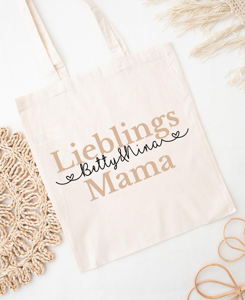May include: Cream-colored tote bag with the words "Lieblings Mama Betty & Nina" printed in a mix of fonts and colors. The bag has two handles and is made of a soft fabric. The design includes heart accents.