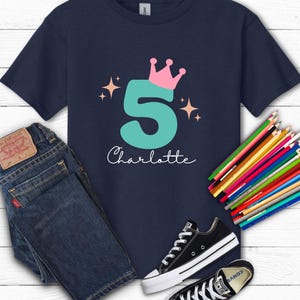 May include: Navy blue t-shirt with a teal number 5 topped with a pink crown and the name "Charlotte" in white script. The shirt is paired with blue jeans, black sneakers, and a set of colored pencils.