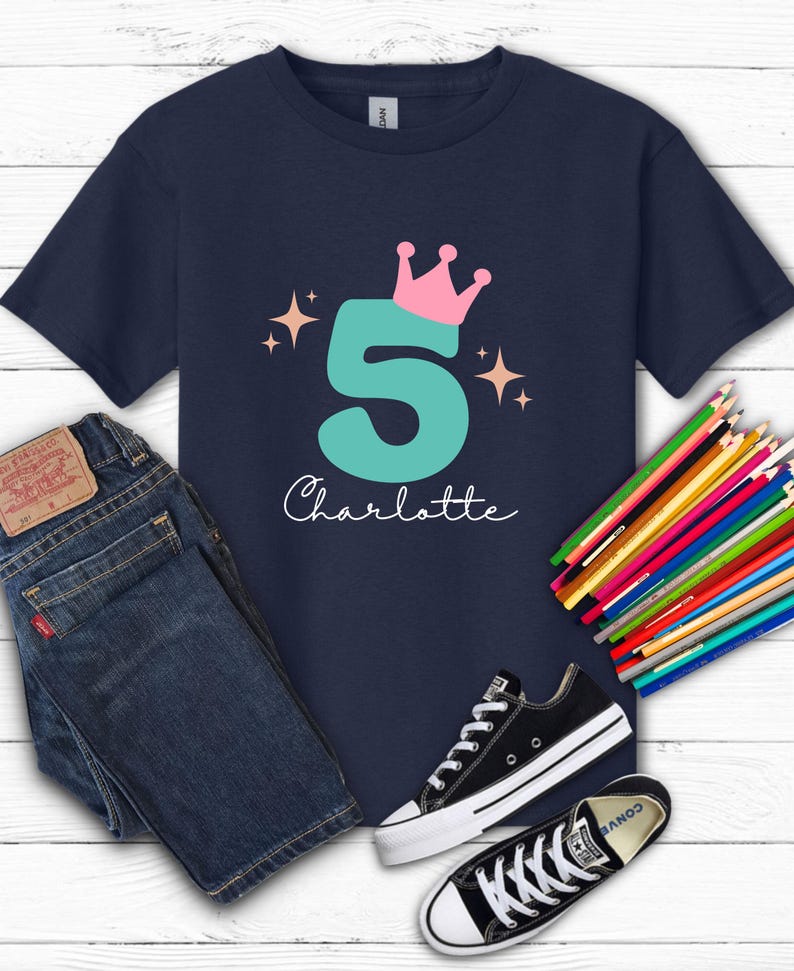 May include: Navy blue t-shirt with a teal number 5 topped with a pink crown and the name "Charlotte" in white script. The shirt is paired with blue jeans, black sneakers, and a set of colored pencils.