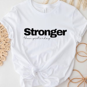May include: White t-shirt with the text "Stronger than yesterday" in black. The shirt is knotted at the waist. The shirt is made of a soft material.