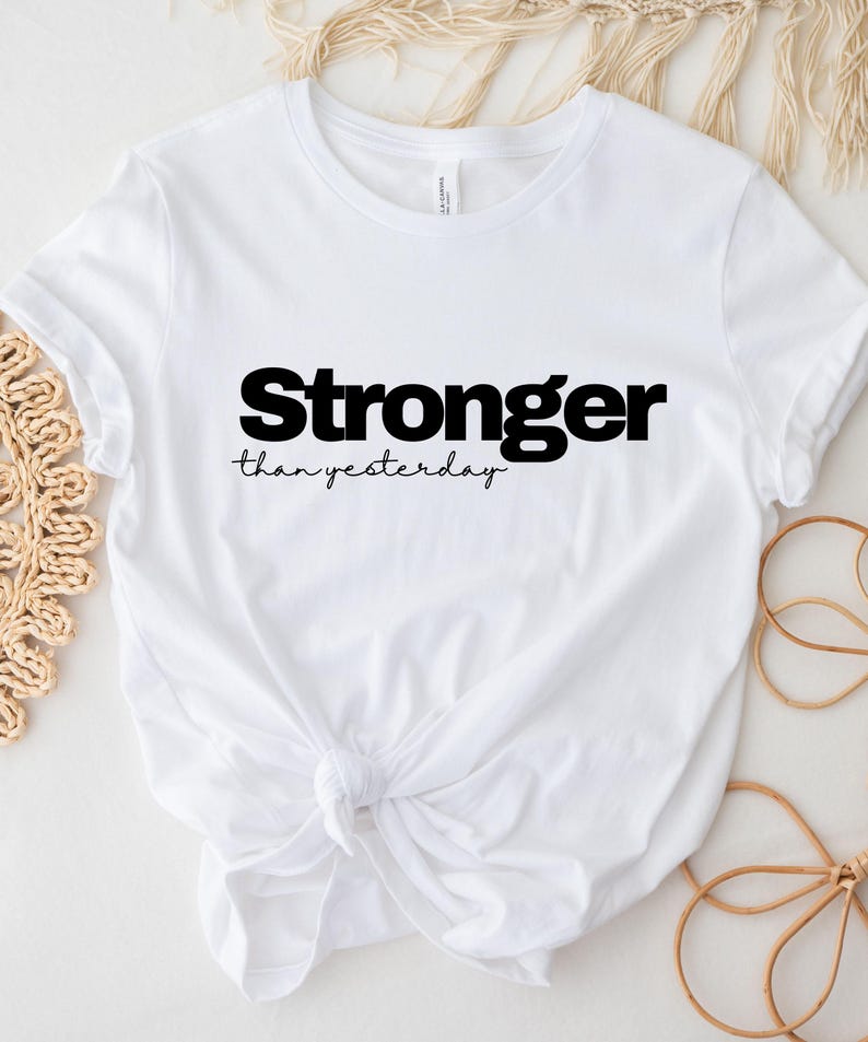 May include: White t-shirt with the text "Stronger than yesterday" in black. The shirt is knotted at the waist. The shirt is made of a soft material.