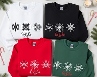 Pode incluir: Quatro camisolas de gola redonda em branco, preto, vermelho e verde escuro. Cada uma apresenta três gráficos de flocos de neve distintos dispostos horizontalmente acima do texto cursivo "hohoho" impresso numa cor contrastante.