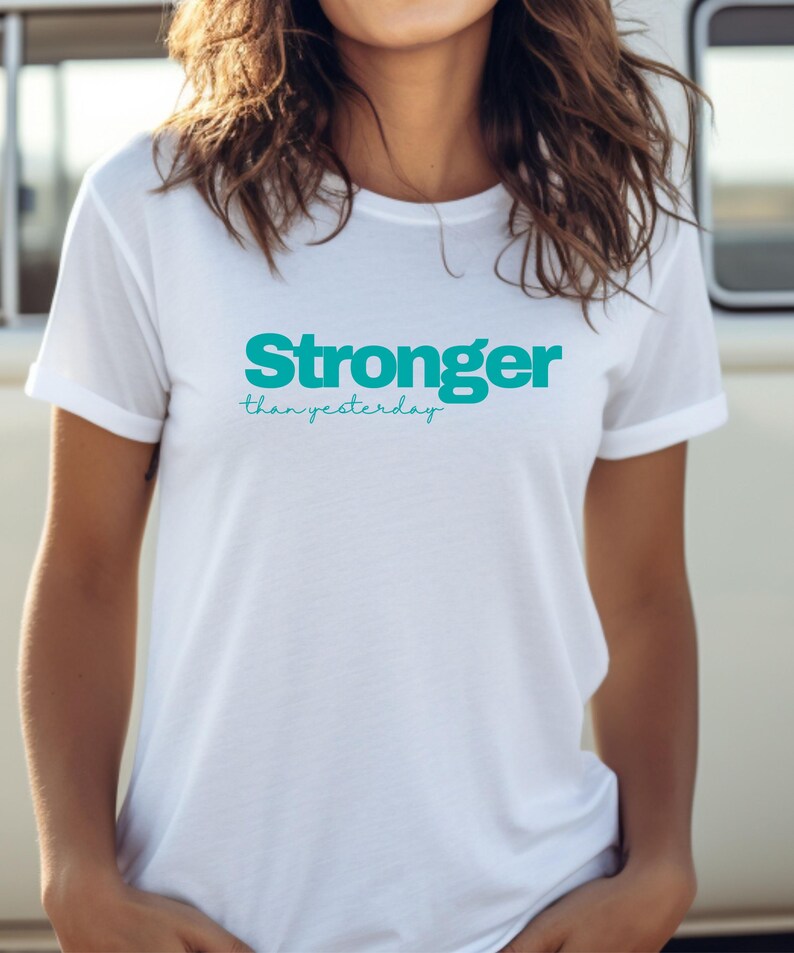 May include: A white t-shirt featuring the teal text "Stronger than yesterday." The shirt has a crew neck and short sleeves. The text is in a bold, sans-serif font with "than yesterday" in a script font.