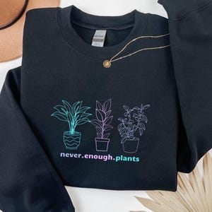 Könnte beinhalten: Schwarzes Sweatshirt mit einem Pflanzenmotiv in Türkis und Lila. Der Text "never.enough.plants" befindet sich unter den Pflanzen. Eine goldene Halskette mit Anhänger liegt über dem Sweatshirt.