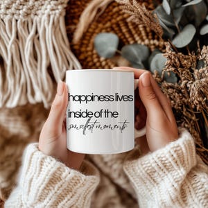 Może przedstawiać: Biały ceramiczny kubek z napisem "happiness lives inside of the smallest moments" czarnym pismem. Kubek trzyma osoba w kremowym swetrze. Tło obejmuje suszone rośliny i tkane tekstury.