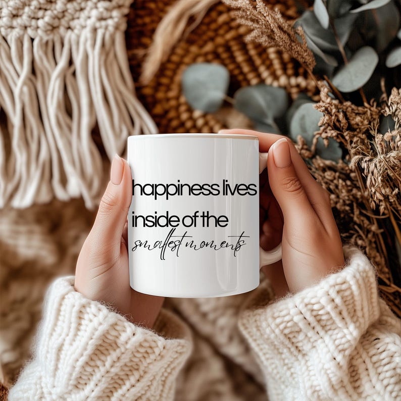 Może przedstawiać: Biały ceramiczny kubek z napisem "happiness lives inside of the smallest moments" czarnym pismem. Kubek trzyma osoba w kremowym swetrze. Tło obejmuje suszone rośliny i tkane tekstury.
