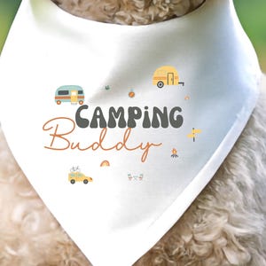 May include: A white triangular pet bandana featuring the text "CAMPING Buddy" in dark gray and orange script. The design includes small colorful illustrations of a camper van, a travel trailer, a yellow car with a bicycle, a campfire, and a rainbow.