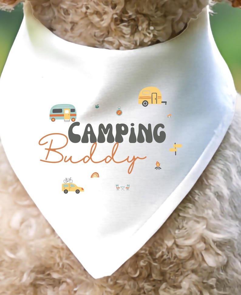 May include: A white triangular pet bandana featuring the text "CAMPING Buddy" in dark gray and orange script. The design includes small colorful illustrations of a camper van, a travel trailer, a yellow car with a bicycle, a campfire, and a rainbow.