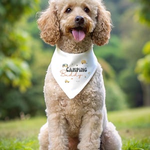 May include: A light-colored curly-haired dog wearing a white triangular pet bandana. The bandana features the text "Camping Buddy" in dark and orange lettering, surrounded by small graphic illustrations of campers, trees, and stars.