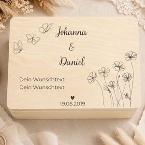 May include: A rectangular light-colored wooden box featuring black line art of three butterflies and a cluster of wildflowers. The text reads Johanna & Daniel, Dein Wunschtext, Dein Wunschtext, and the date 19.06.2019 with a small heart symbol.