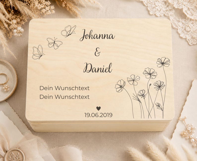May include: A rectangular light-colored wooden box featuring black line art of three butterflies and a cluster of wildflowers. The text reads Johanna & Daniel, Dein Wunschtext, Dein Wunschtext, and the date 19.06.2019 with a small heart symbol.