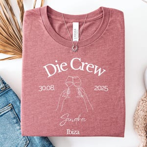 May include: A folded mauve t-shirt with the text "Die Crew" and the date "30.08.2025" printed in white. Below the text is a line drawing of two wine glasses clinking, with the name "Sandra" and the word "Ibiza" underneath. A silver necklace with a sun pendant is on the shirt.