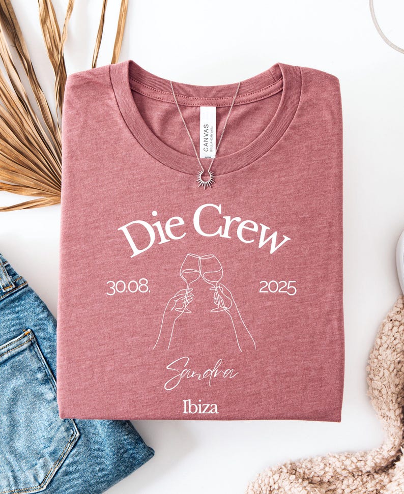 May include: A folded mauve t-shirt with the text "Die Crew" and the date "30.08.2025" printed in white. Below the text is a line drawing of two wine glasses clinking, with the name "Sandra" and the word "Ibiza" underneath. A silver necklace with a sun pendant is on the shirt.