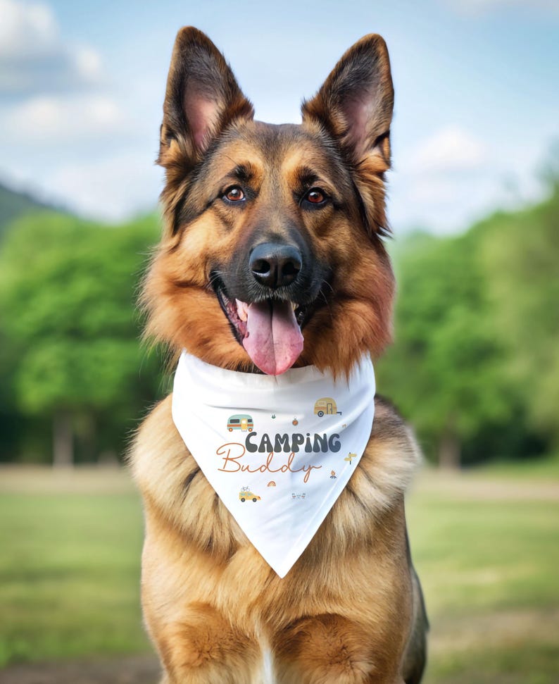 May include: A white triangular dog bandana featuring the text "Camping Buddy" in dark gray and orange script. The fabric is decorated with small illustrations of campers, pine trees, and mountains in muted earth tones, worn by a German Shepherd.