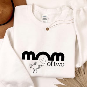 Może przedstawiać: Biała bluza z czarnym napisem "mom of two" ze znakiem serca. Poniżej napisane kursywą imiona "Denise" i "Angelika". Złoty naszyjnik i brązowy kapelusz w tle.