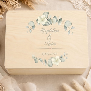 May include: A rectangular light-colored wooden box featuring a printed design of eucalyptus leaves and white flowers arranged in a wreath. The text "Magdalena & Anton" is centered, followed by a small heart and the date "15.07.2026" below.