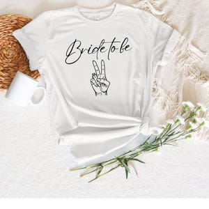 May include: White t-shirt with the text "Bride to be" in a cursive font and a black illustration of a hand making a peace sign. The shirt is knotted at the waist and is on a white surface with a white mug and a woven basket.