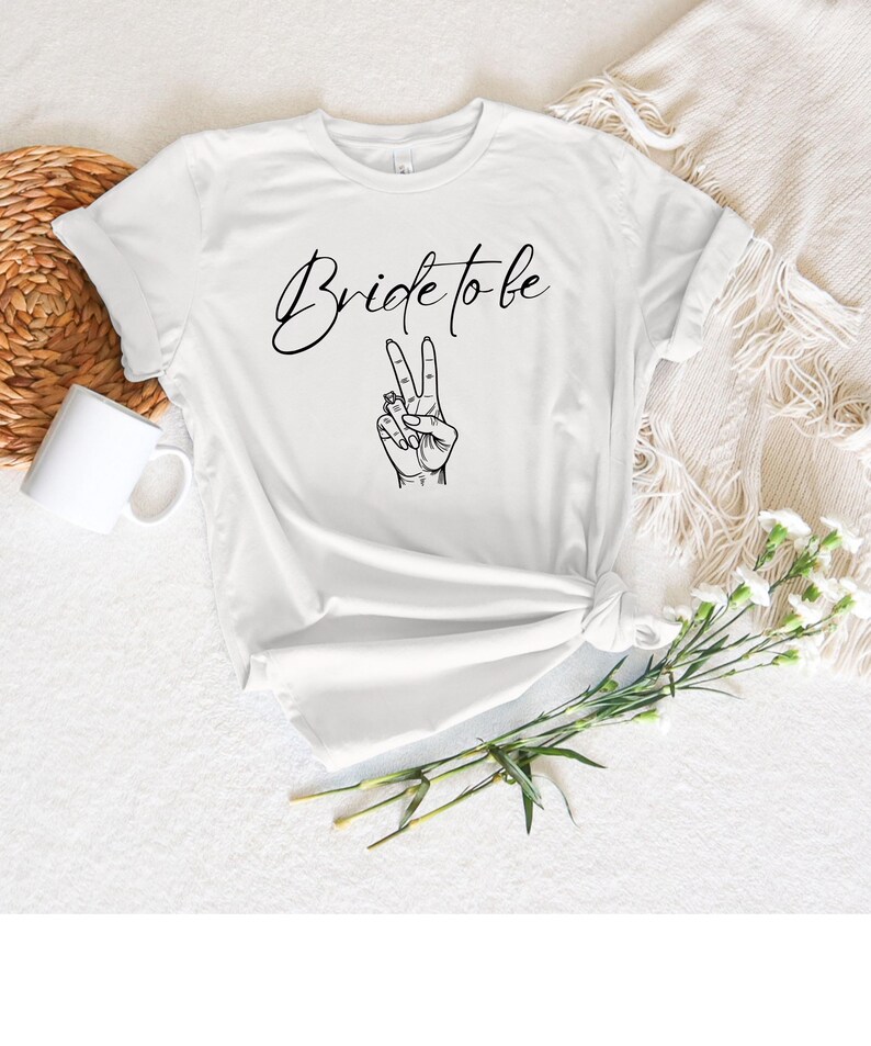 May include: White t-shirt with the text "Bride to be" in a cursive font and a black illustration of a hand making a peace sign. The shirt is knotted at the waist and is on a white surface with a white mug and a woven basket.
