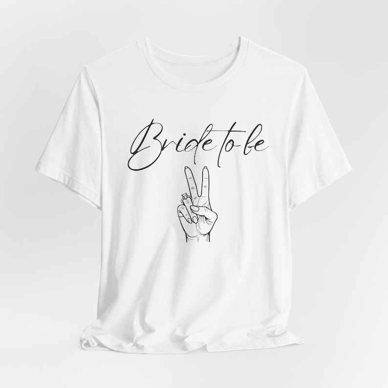 May include: White t-shirt with the text "Bride to be" in a cursive font. Below the text is a black and white illustration of a hand making a peace sign, with a ring on the ring finger.