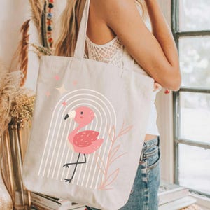 May include: Beige canvas tote bag featuring a pink flamingo design. The flamingo is inside a white arched design with vertical lines. Pink stars and a pink leaf design are also present. The bag has two beige straps.