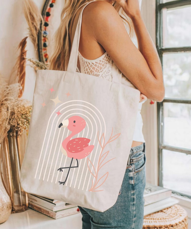 May include: Beige canvas tote bag featuring a pink flamingo design. The flamingo is inside a white arched design with vertical lines. Pink stars and a pink leaf design are also present. The bag has two beige straps.