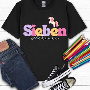 K&ouml;nnte beinhalten: Schwarzes T-Shirt mit dem Namen "Sieben" in Lila, Gelb und Rosa und dem Namen "Melanie" in wei&szlig;er Schrift. Ein Cartoon-Einhorn befindet sich &uuml;ber dem Namen. Das Shirt wird mit blauen Jeans und schwarzen Turnschuhen kombiniert.