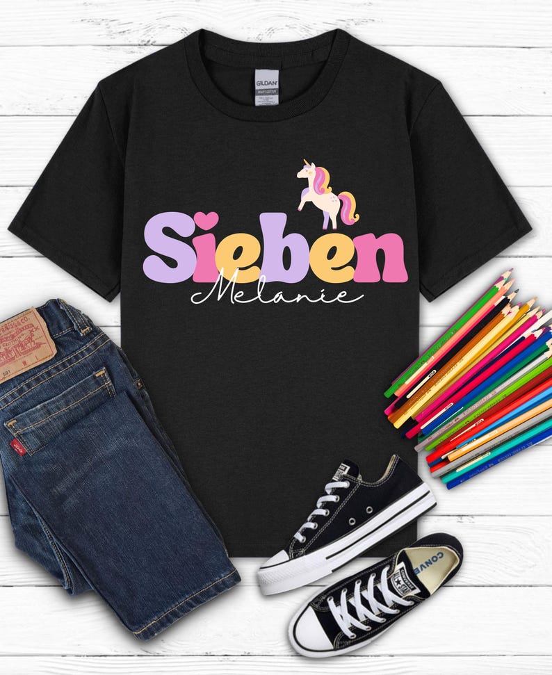 K&ouml;nnte beinhalten: Schwarzes T-Shirt mit dem Namen "Sieben" in Lila, Gelb und Rosa und dem Namen "Melanie" in wei&szlig;er Schrift. Ein Cartoon-Einhorn befindet sich &uuml;ber dem Namen. Das Shirt wird mit blauen Jeans und schwarzen Turnschuhen kombiniert.