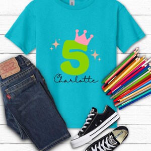 May include: A turquoise t-shirt with a large green number 5, a pink crown, and the name "Charlotte" in black script. The shirt is paired with blue jeans, black Converse sneakers, and a set of colored pencils.