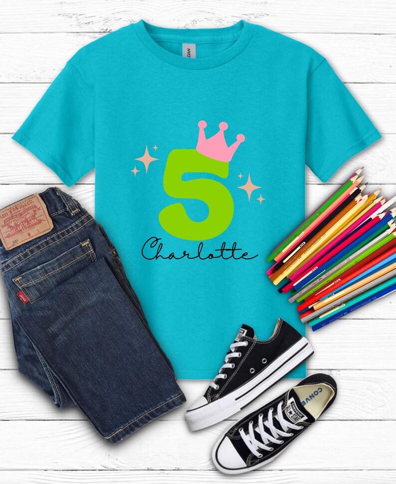 May include: A turquoise t-shirt with a large green number 5, a pink crown, and the name "Charlotte" in black script. The shirt is paired with blue jeans, black Converse sneakers, and a set of colored pencils.