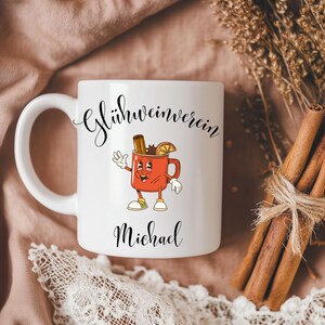 Pu&ograve; includere: Tazza in ceramica bianca con un'illustrazione a cartoni animati di una tazza rossa con un bastoncino di cannella, anice stellato e una fetta d'arancia. La tazza ha il testo "Gl&uuml;hweinverein" e "Michael" in corsivo nero.