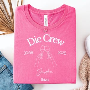 May include: A hot pink t-shirt with the words "Die Crew" in white script. Below the text is a line drawing of two hands holding wine glasses, the date 30.08.2025, the name "Sandra", and the word "Ibiza". A silver necklace with a sun pendant is on the shirt.