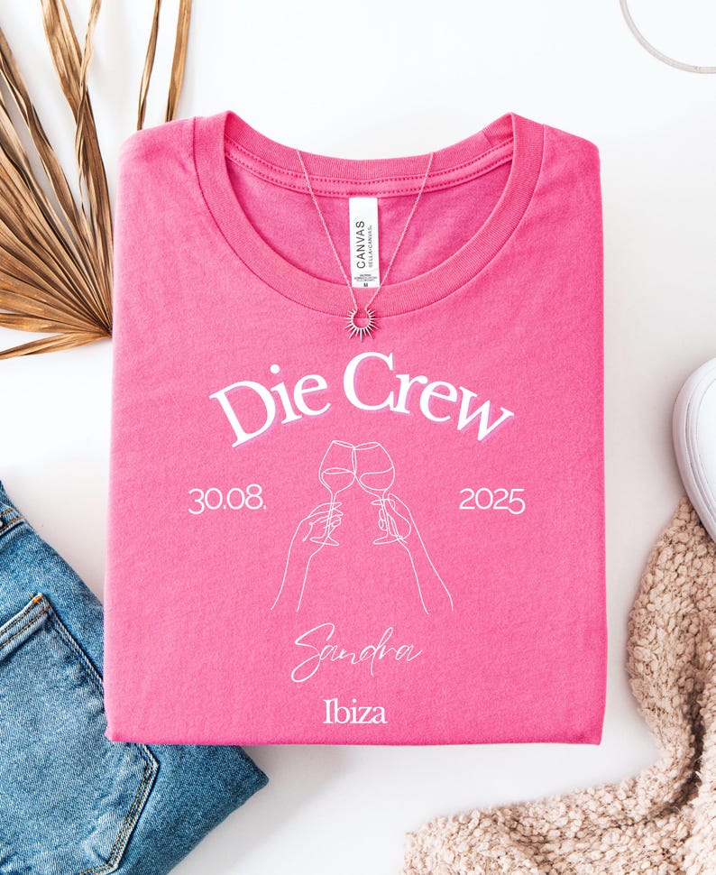 May include: A hot pink t-shirt with the words "Die Crew" in white script. Below the text is a line drawing of two hands holding wine glasses, the date 30.08.2025, the name "Sandra", and the word "Ibiza". A silver necklace with a sun pendant is on the shirt.