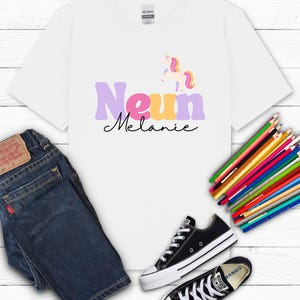 K&ouml;nnte beinhalten: Wei&szlig;es T-Shirt mit dem Namen "Melanie" und einem Einhorn-Motiv. Das Shirt wird mit blauen Jeans und schwarzen Converse-Sneakern kombiniert. Ein B&uuml;ndel Buntstifte ist ebenfalls zu sehen.