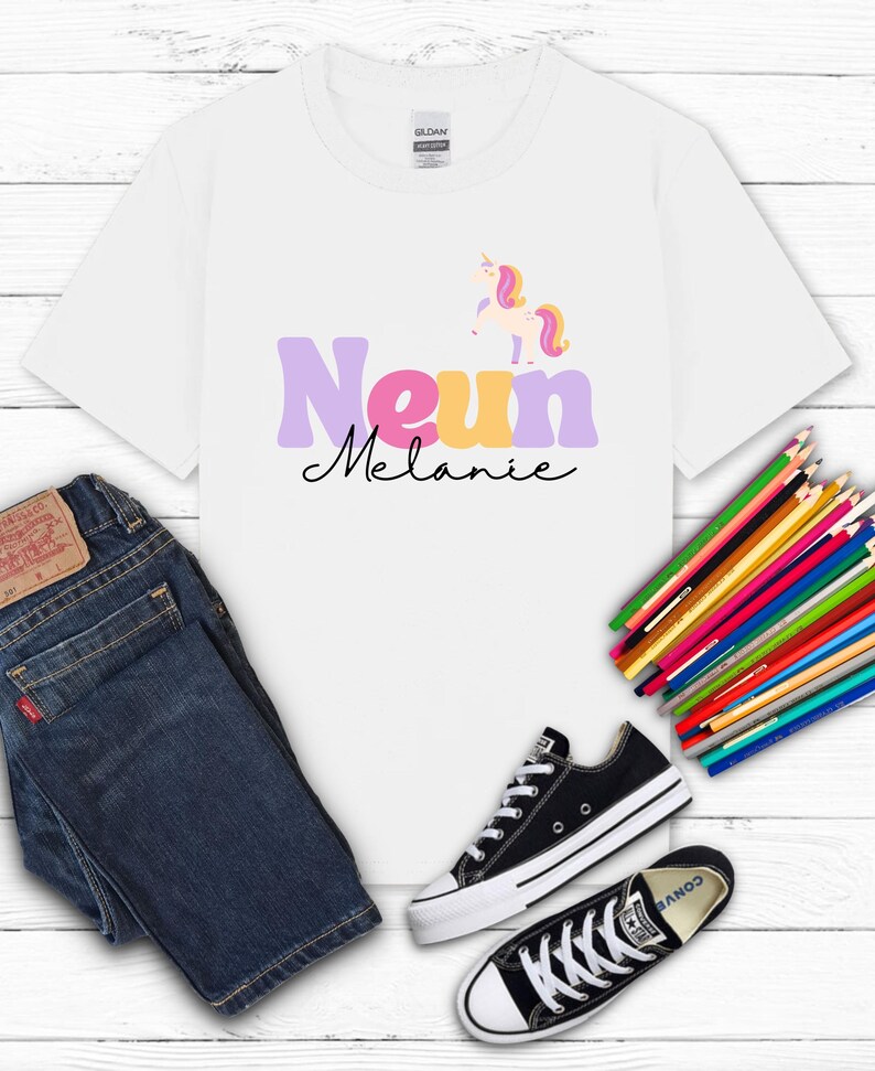 K&ouml;nnte beinhalten: Wei&szlig;es T-Shirt mit dem Namen "Melanie" und einem Einhorn-Motiv. Das Shirt wird mit blauen Jeans und schwarzen Converse-Sneakern kombiniert. Ein B&uuml;ndel Buntstifte ist ebenfalls zu sehen.