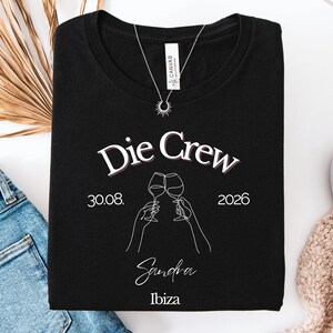 May include: A black t-shirt featuring the white text Die Crew, the date 30.08. 2026, the name Sandra, and the word Ibiza. The graphic includes a white line art illustration of two hands holding wine glasses in a toast, centered on the chest.